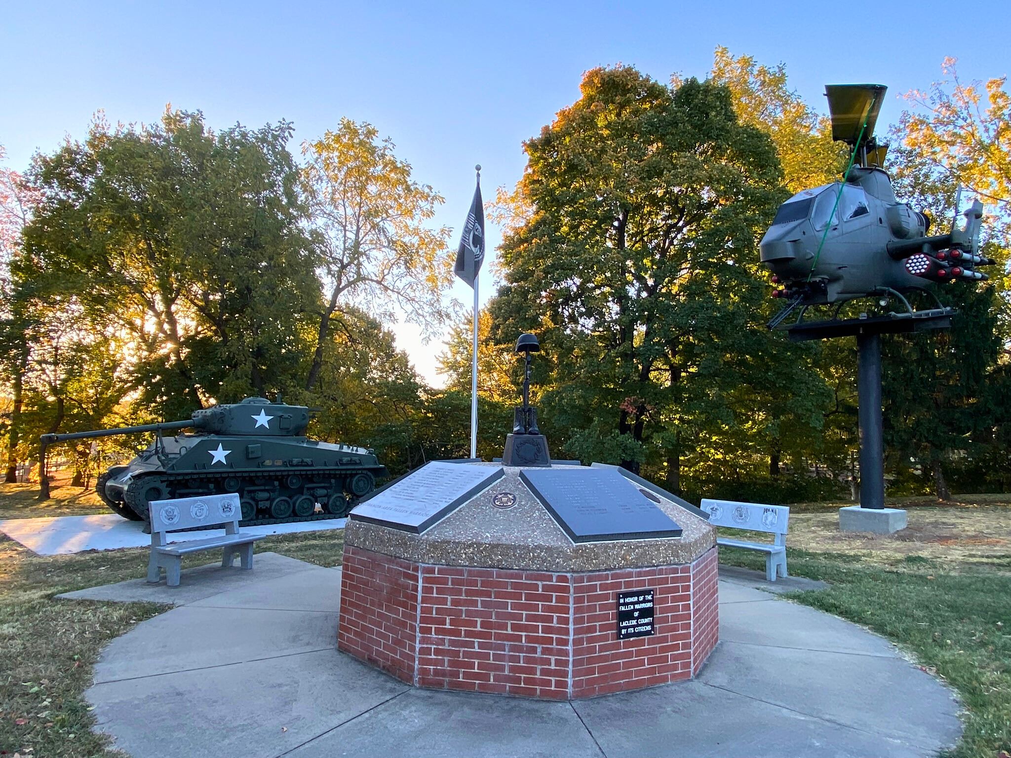 Brick memorial with plaques and benches at Lebanon-Area Veterans Memorial Park, with a Sherman tank on the left and an AH‑1 Cobra helicopter on display to the right among trees.