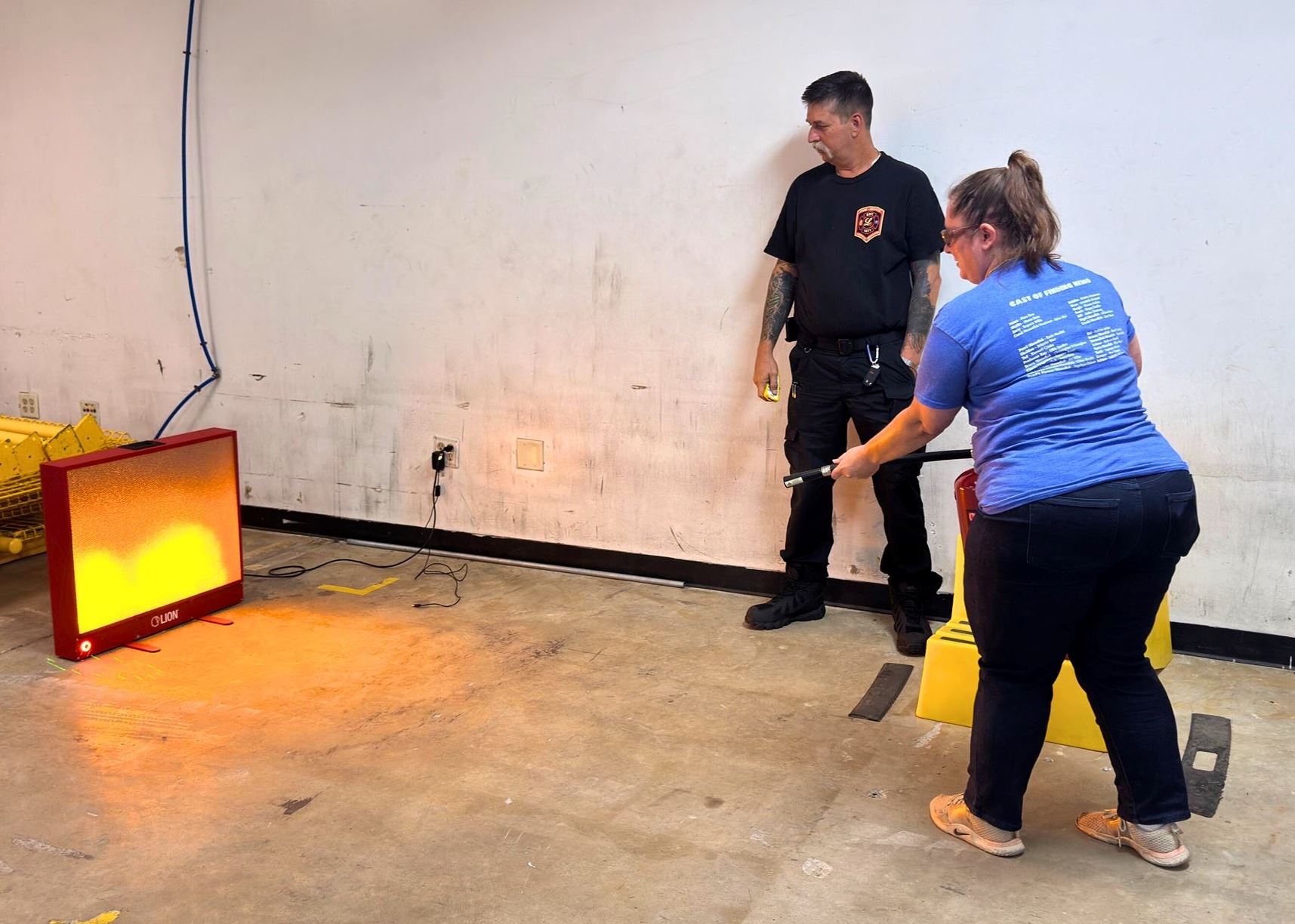 A woman practices using a fire extinguisher on a digital fire  firefighter looking on.